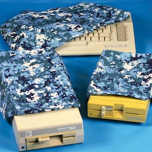 May include: A Commodore 64 computer and disk drives are covered with blue and white digital camouflage fabric covers. The keyboard is visible under a cover. The disk drives are beige and yellow.