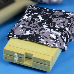 Peut inclure: Un ordinateur vintage beige avec une housse de camouflage numérique noire, blanche et grise. L'ordinateur possède un lecteur de disquette et un interrupteur blanc. La housse est drapée sur le dessus de l'ordinateur.