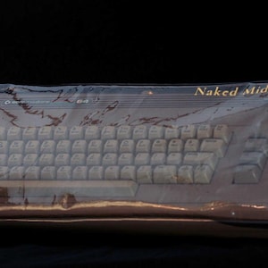 May include: A Commodore 64 computer keyboard encased in a clear, crinkled plastic protective cover. The keyboard features light-colored keys and a dark chassis. The text 'Naked Midnight' is printed in gold lettering on the upper right corner.