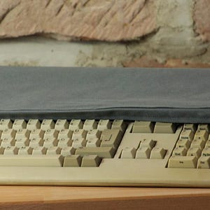 Peut inclure: Un clavier d'ordinateur beige avec une housse grise. Le clavier comporte de nombreuses touches et est incliné vers le haut. La housse est drapée sur le dessus du clavier, le protégeant de la poussière et des débris.