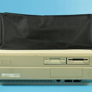 Peut inclure: Un ordinateur Commodore Amiga 2000 vintage, recouvert d'une housse noire. L'ordinateur est beige avec deux lecteurs de disquettes et une étiquette indiquant "Commodore AMIGA 2000". Le fond est bleu uni.
