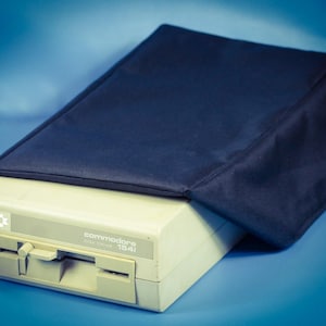 May include: A vintage Commodore 1541 disk drive, beige in color, with a dark blue cover. The drive has a disk slot and a power button. The text "Commodore disk drive 1541" is printed on the front.