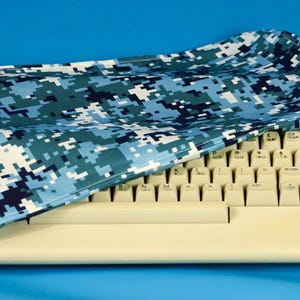 May include: A beige Commodore 64 personal computer keyboard with a blue and white digital camouflage cover. The keyboard has white keys and gray function keys. The cover is draped over the keyboard.