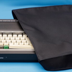 Pode incluir: Um computador doméstico Toshiba HX-10 com chassis cinza-escuro e teclado cinza-claro com teclas brancas, uma tecla verde e teclas de função cinza-escuras. O texto "TOSHIBA HOME COMPUTER HX-10" e "MADE IN JAPAN" é visível. Uma capa de tecido preta está incluída.