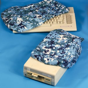 May include: Two computer covers with a blue and white digital camouflage pattern. One cover is on a keyboard, the other on a beige floppy disk drive. The background is a solid blue.