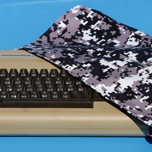 Peut inclure: Un ordinateur Commodore 64 beige avec un clavier noir est partiellement recouvert d'un tissu de camouflage numérique. Le clavier a des touches noires avec des lettres blanches. Le tissu présente un motif de pixels noirs, blancs et gris.