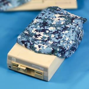 Könnte beinhalten: Ein beigefarbener Commodore 128 Computer mit einer blau-weiß getarnten Hülle. Die Hülle ist über die Oberseite des Computers drapiert. Der Computer hat ein Diskettenlaufwerk und einen Einschaltknopf.