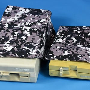 Peut inclure: Deux lecteurs de disquettes Commodore 1541 vintage, un beige et un jaune, sont recouverts d'un tissu à motif camouflage pixélisé noir, blanc et gris. Les lecteurs sont sur un fond bleu.