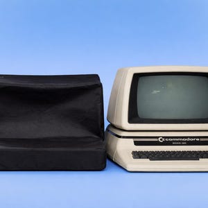 May include: A vintage Commodore 64 computer with a cream-colored body and black keyboard, next to a black cover. The computer has a screen and the Commodore logo. The model number is visible.