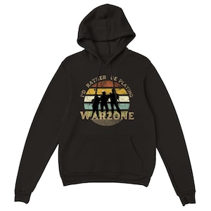 Könnte beinhalten: Schwarzer Hoodie mit einer Grafik im Vintage-Stil von drei Personen, die Warzone spielen. Die Grafik zeigt einen Sonnenuntergang mit einem Farbschema aus Gelb, Orange und Rot. Der Text lautet "I'd rather be playing Warzone".