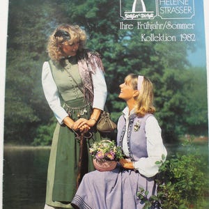 May include: Two women wearing traditional dirndl dresses. The woman on the left is wearing a green dirndl with a white blouse and a brown shawl. The woman on the right is wearing a purple dirndl with a white blouse and a blue waistcoat. Both women are holding flowers and smiling. The image is from a fashion catalogue for "Schläger Dirndl" and "Creation Helene Strasser" with the text "Ihre Frühjahr/Sommer Kollektion 1982".
