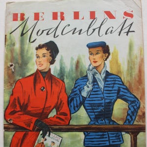 Pode incluir: Capa de revista vintage "BERLINS Modenblatt" com duas mulheres em casacos. Uma usa um casaco vermelho e luvas pretas, segurando uma revista. A outra, um fato azul às riscas. Texto alemão e estilo retro.