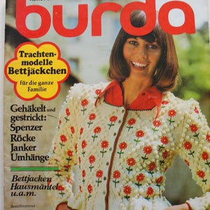 May include: A vintage magazine cover showing a woman in a cream cardigan with red floral embroidery. The magazine title "burda" is in large red letters. The cover includes German text promoting knitting and crochet patterns for traditional designs.