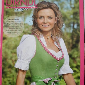 Könnte beinhalten: Ein Magazin-Cover mit dem Titel Dirndl Revue 2011, das ein grünes Dirndl mit weißer Bluse, rosa Blumenborte und passender Schürze zeigt. Der Text enthält Informationen zu Schnittmustern für die Größen 32 bis 50 sowie Kindergrößen 92 bis 164.