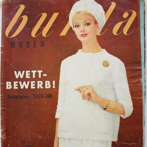 Könnte beinhalten: Ein vintage Burda Moden Magazin-Cover vom Oktober 1960 mit einer Person in einer weißen, strukturierten Jacke, passendem Rock und weißer Pillbox-Mütze. Das Cover enthält den Text WETTBEWERB! Hauptpreis: 5000 DM und 10/1960 OKTOBER.