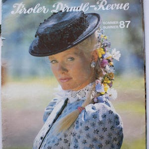 May include: A vintage magazine cover titled Tiroler Dirndl-Revue Sommer 87 featuring a person wearing a light blue floral patterned dress with a white lace collar, a black hat, and a braided hairstyle adorned with colourful small flowers.