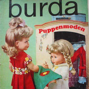 Könnte beinhalten: Titelseite eines Burda-Magazins aus der Vintage-Zeit mit zwei Puppen. Eine Puppe trägt ein rotes Kleid mit floralen Details, die andere ein weißes Kleid. Der Magazintitel "burda" steht in großen schwarzen Buchstaben.