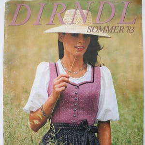 May include: A vintage magazine cover titled DIRNDL SOMMER '83 featuring a person wearing a straw hat, a white blouse with puffed sleeves, and a pink bodice with white dots over a dark patterned skirt. The text includes 39 Modelle mit Schnittmusterbogen.