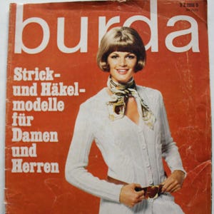 May include: A vintage magazine cover with the word "burda" in large white letters on a red background. The cover features a woman wearing a white knit cardigan and skirt. Text on the cover reads "Strick- und Häkelmodelle für Damen und Herren."