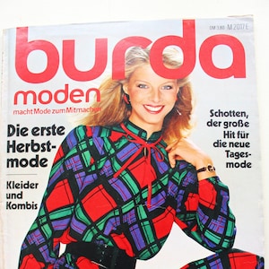 Può includere: Copertina della rivista vintage "burda moden" dell'agosto 1980. Mostra una donna con un abito a quadri rossi, verdi e viola. Il titolo della rivista è rosso. Il testo include "Die erste Herbst-mode" e "August 1980".