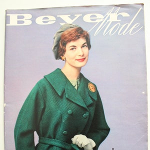 May include: A green double-breasted jacket with a belt and white gloves. The model is wearing a light blue hat and has short brown hair. The magazine cover reads "Beyer Mode" and "35. JAHRGANG".