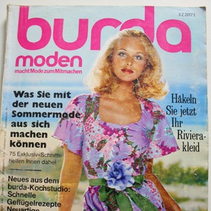 May include: A magazine cover featuring a woman wearing a floral dress with a green sash and a blue flower brooch. The magazine is titled "burda moden" and the text "macht Mode zum Mitmachen" is below the title. The cover also features the text "Was Sie mit der neuen Sommermode aus sich machen können" and "75 Exklusiv-Schnitte helfen Ihnen dabei".