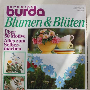Peut inclure: Une couverture de magazine présentant un motif floral avec le titre "Blumen & Blüten" et le texte "Über 50 Motive Alles zum Selber-machen". La couverture comprend également un gros plan d'un motif de broderie florale et le texte "EXTRA: Multi-Bügelmuster E 264".