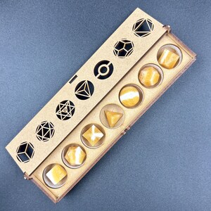 May include: A wooden box with a sliding lid, containing seven yellow-orange gemstone dice. The lid features geometric cutouts. The dice are in various shapes, including cubes, spheres, and pyramids. The box is on a gray surface.