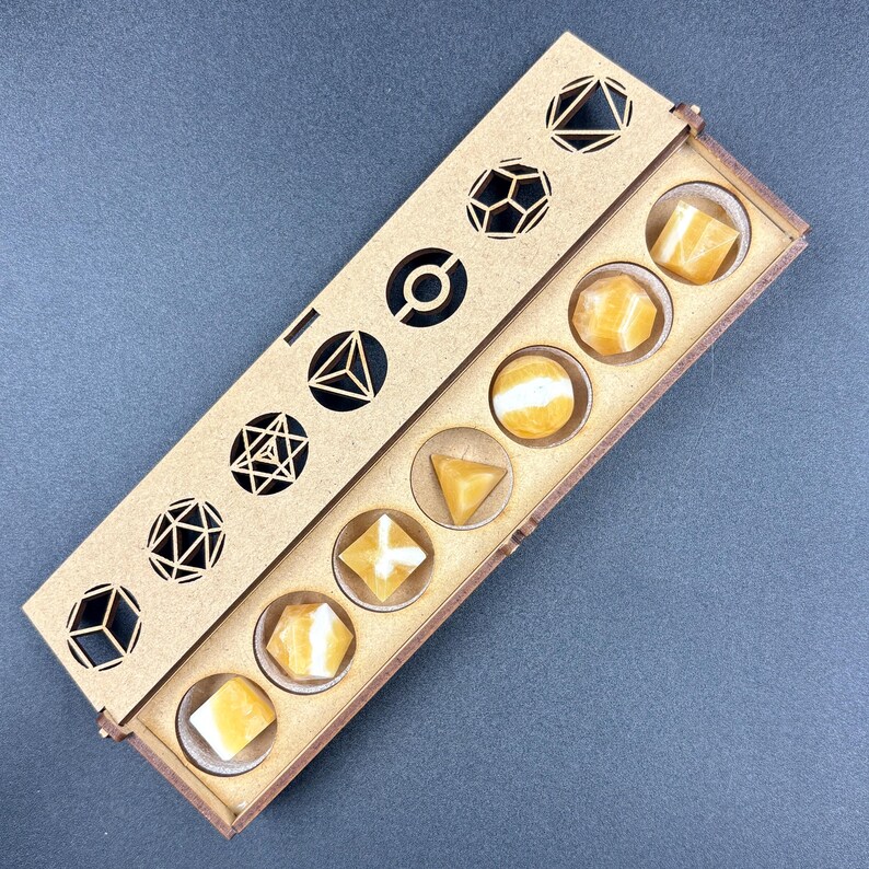 May include: A wooden box with a sliding lid, containing seven yellow-orange gemstone dice. The lid features geometric cutouts. The dice are in various shapes, including cubes, spheres, and pyramids. The box is on a gray surface.