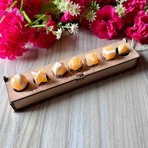 May include: A wooden box with six orange and white SANA AGATE crystals in various shapes. The crystals include a sphere, a star, and geometric shapes. The box is on a wooden surface, with pink flowers in the background.