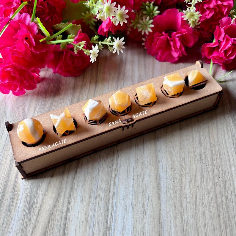 May include: A wooden box with six orange and white SANA AGATE crystals in various shapes. The crystals include a sphere, a star, and geometric shapes. The box is on a wooden surface, with pink flowers in the background.