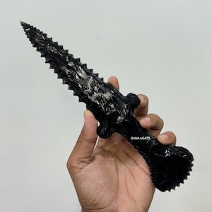 May include: A black obsidian dagger with a serrated blade and a textured handle. The dagger is held in a hand against a white background. The text "SANA AGATE" is visible on the handle.