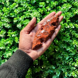 May include: A red agate arrowhead with a jagged edge and a smooth, polished surface. The arrowhead is about 5 inches long and has a natural, organic shape.