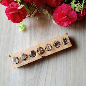 May include: A wooden display featuring seven clear crystal stones in various geometric shapes. The display is labeled "SANA AGATE" and is set against a backdrop of red flowers. The crystals are faceted and appear to be quartz or similar gemstones.