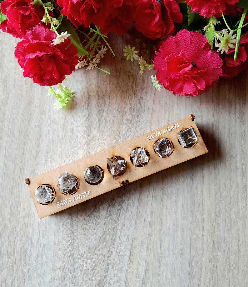May include: A wooden display featuring seven clear crystal stones in various geometric shapes. The display is labeled "SANA AGATE" and is set against a backdrop of red flowers. The crystals are faceted and appear to be quartz or similar gemstones.