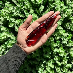 May include: A red agate arrowhead with a smooth, polished surface. The arrowhead is held in a hand against a green leafy background. The text "SANA AGATE" is visible on the arrowhead.
