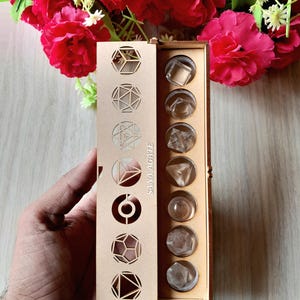 May include: A wooden box with geometric cutouts and the text "SANA AGATE" holds clear, faceted crystals. The box is open, revealing the crystals in individual compartments. Red flowers are in the background.