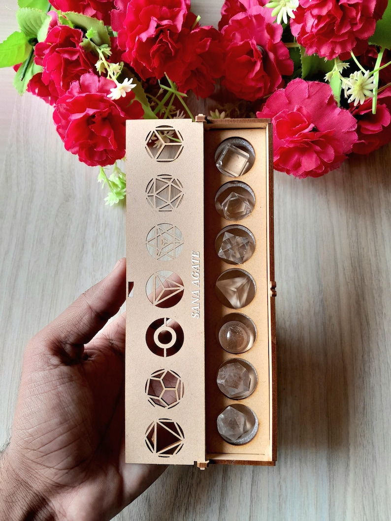 May include: A wooden box with geometric cutouts and the text "SANA AGATE" holds clear, faceted crystals. The box is open, revealing the crystals in individual compartments. Red flowers are in the background.