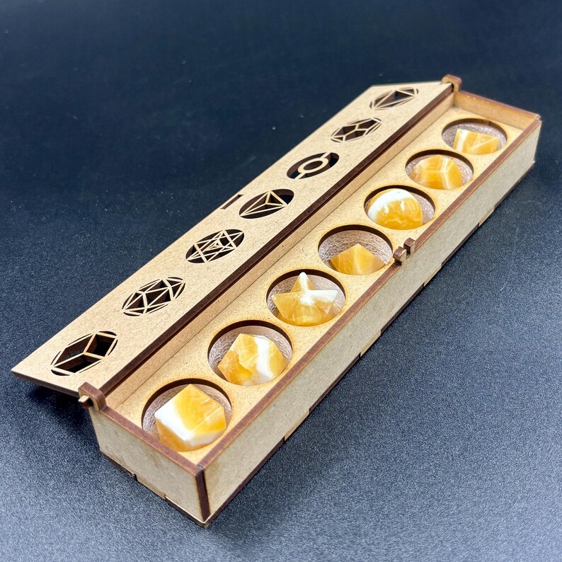 May include: A wooden box with a hinged lid, containing seven orange and white gemstones. The lid features geometric cutouts. The gemstones are round and fit into individual circular compartments within the box.