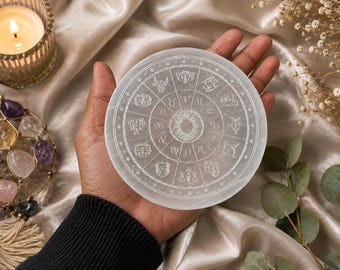 May include: A circular white selenite charging plate featuring an engraved zodiac wheel with twelve astrological symbols arranged in a radial pattern around a central sunburst design, with a smooth, translucent, and polished stone surface.