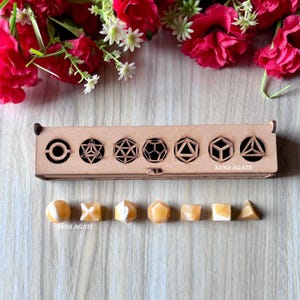 May include: A wooden box with geometric cutouts and the text "SANA AGATE" is displayed with a set of orange and white geometric crystals. The crystals include spheres, cubes, and pyramids. Red flowers and greenery are in the background.