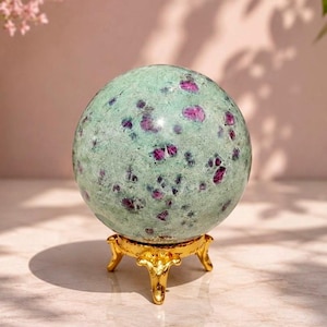 May include: A polished spherical stone featuring a pale mint green base with scattered deep magenta and purple inclusions throughout the surface. The sphere rests securely on a small, ornate, reflective gold-toned metal tripod display stand.
