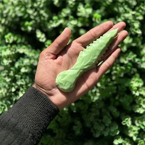 Puede incluir: Una punta de flecha de piedra verde en forma de hoja con un borde dentado. La punta de flecha se sostiene en una mano.