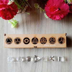May include: A wooden box with geometric cutouts and clear crystal shapes. The box has various geometric designs. Below the box are several clear crystal shapes, including spheres, pyramids, and cubes. The text "SANA AGATE" is visible.