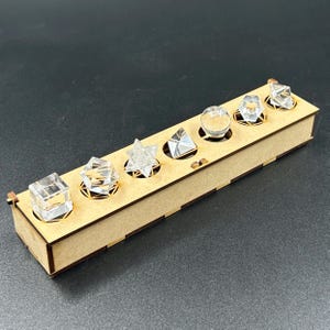 May include: A wooden box with six clear, geometric shapes: a cube, a diamond, a star, a pyramid, a sphere, and a multifaceted shape. The shapes are set in circular cutouts. The box is made of light brown wood.