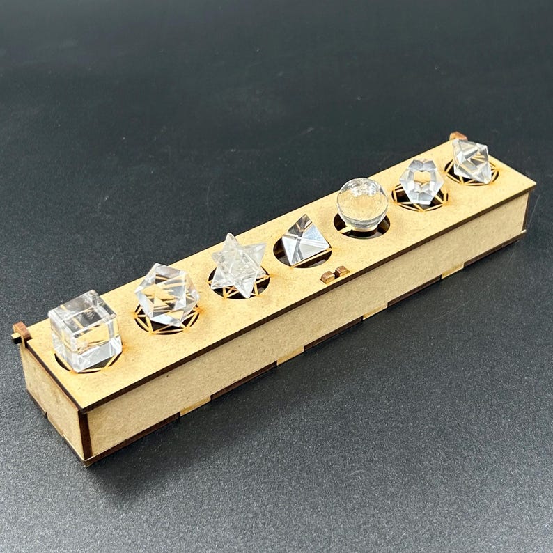 May include: A wooden box with six clear, geometric shapes: a cube, a diamond, a star, a pyramid, a sphere, and a multifaceted shape. The shapes are set in circular cutouts. The box is made of light brown wood.