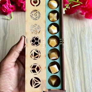 May include: A wooden box with geometric cutouts on the lid, containing various shaped yellow agate stones. The box is open, revealing the stones in a teal-colored compartment. The text "SANA AGATE" is printed on the side of the box.
