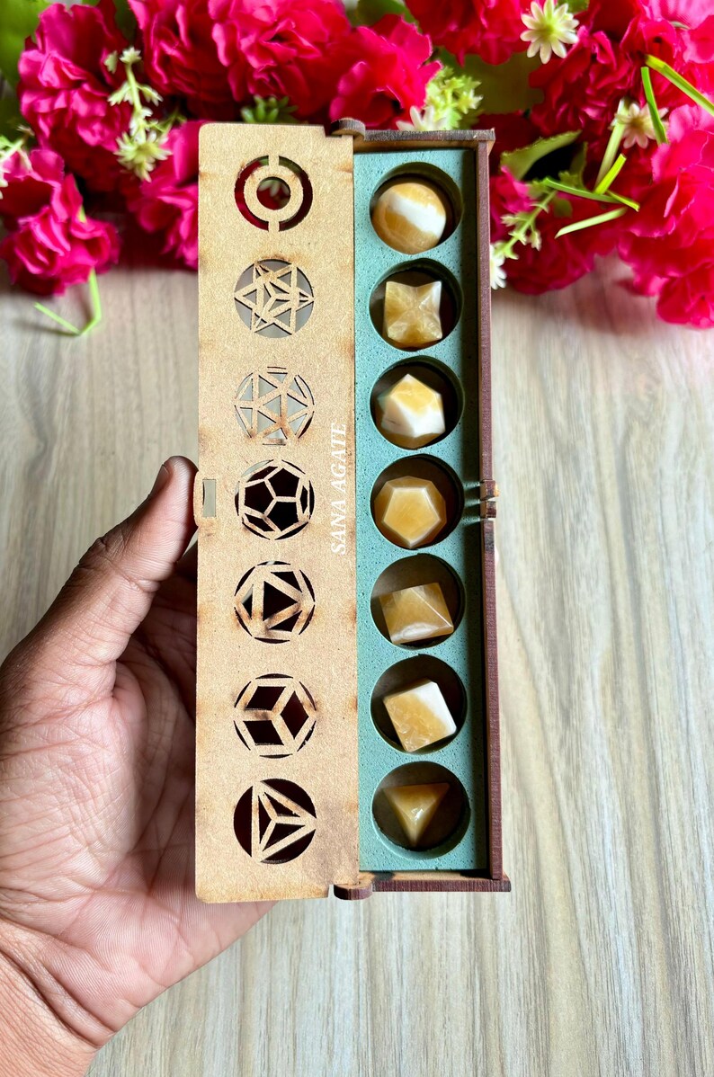 May include: A wooden box with geometric cutouts on the lid, containing various shaped yellow agate stones. The box is open, revealing the stones in a teal-colored compartment. The text "SANA AGATE" is printed on the side of the box.