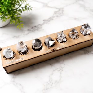 May include: A wooden display box with six clear crystal formations. The crystals are various shapes, including a sphere, pyramids, and a cube. The box has the text "SANA AGATE" on it. A small green plant in a white pot is in the background.