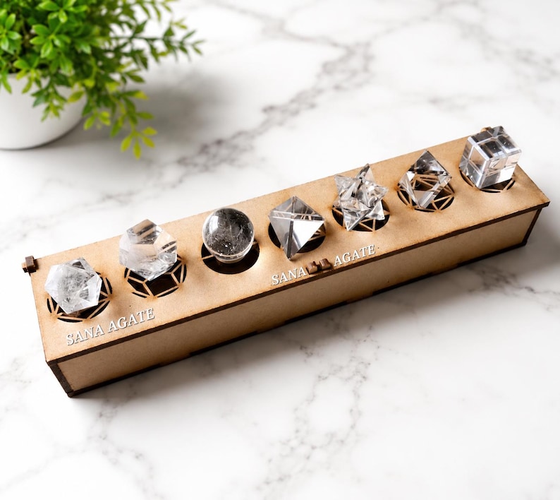 May include: A wooden display box with six clear crystal formations. The crystals are various shapes, including a sphere, pyramids, and a cube. The box has the text "SANA AGATE" on it. A small green plant in a white pot is in the background.